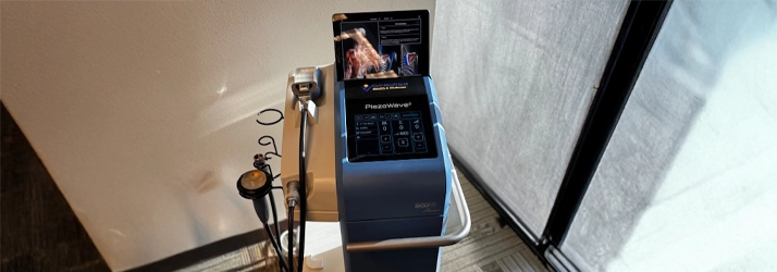 A medical device called PiezoWave is shown on a wheeled stand. It has a control panel with buttons and a display screen showing anatomical images. Several connected accessories, including a handheld applicator, are attached to the unit.