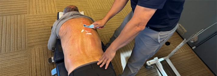 A person is lying face down on a treatment table while another individual applies a handheld device to the person's back. The procedure appears to involve targeted therapy on the back area, with adhesive markers placed on the skin.