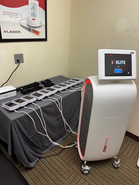 A medical device with a touchscreen labeled “i ELITE” is positioned next to a table holding multiple connected pads, likely used for laser or light therapy treatments.