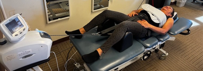 A person is lying on a specialized traction table with straps around the waist and legs, connected to a machine used for spinal decompression therapy.